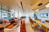 Located in Washington, DC's vibrant NoMa neighborhood, this elevated amenity lounge offers a thoughtfully designed open-plan space that seamlessly blends social, lounge, and collaborative areas. The interior is anchored by a striking warm-toned wood plank ceiling that runs the length of the room, lending a natural, organic quality to the contemporary design. Floor-to-ceiling windows wrap the perimeter, flooding the space with abundant natural light and framing sweeping panoramic views of the Washington, DC skyline and surrounding cityscape.

The layout is organized into distinct yet fluid zones. A generous communal work and dining area features long natural wood tables paired with leather and wood chairs beneath a sculptural pendant lighting track. A lounge zone offers plush velvet seating, curved sofas, and vintage-inspired armchairs arranged over richly patterned area rugs, creating an inviting, residential atmosphere. A sleek dark metal shelving unit serves as a refined room divider, displaying curated ceramics and decorative objects.

Lush indoor plants are distributed throughout, enhancing the biophilic character of the space. A built-in wet bar with teal cabinetry and a convex mirror accent adds a functional yet stylish touch. Large-format light stone tile flooring ties the entire space together with a clean, understated elegance.