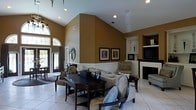 Located at 560 New Hope Rd in Goldsboro, North Carolina, this single-story property offers a sophisticated and well-appointed interior designed with both style and comfort in mind. The main living area immediately draws attention with its dramatic vaulted ceilings that soar overhead, creating a grand sense of openness and volume. Warm golden-tan walls are complemented by crisp white wainscoting and trim, lending the space a classic, refined aesthetic. Recessed lighting is thoughtfully placed throughout, ensuring the interior is well-lit and inviting. Large arched windows and glass-paneled entry doors allow natural light to pour in, further brightening the space and offering views to the outdoors. The living area is elegantly furnished with a neutral sofa, coordinating upholstered chairs in soft blue tones, and a dark wood dining or game table with matching chairs, creating a versatile and functional gathering space. Built-in shelving and niche displays add decorative character, while a fireplace with a white mantle serves as a refined focal point. Light-toned tile flooring runs throughout, tying the design elements together seamlessly. Framed artwork on the walls and carefully curated décor complete the polished, welcoming ambiance of this distinctive property.