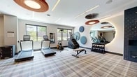 This thoughtfully designed fitness center offers a generously sized, single-floor layout that accommodates a comprehensive range of workout equipment. The space is divided into distinct functional zones, each catering to different exercise disciplines. The main cardio area features multiple treadmills, an elliptical trainer, a stair climber, and a stationary bike, all positioned to take advantage of the natural light streaming in through large windows that offer views of surrounding greenery. A dedicated free weights section includes a full rack of dumbbells alongside a flat workout bench, while a functional trainer cable machine anchors a separate strength training area. The interior design is notably contemporary, with a neutral-toned plaid carpet adding warmth and texture underfoot. Eye-catching circular acoustic panels in shades of blue and terracotta are mounted on the walls, serving both a decorative and functional purpose. The ceiling features a striking combination of large sculptural ring pendant lights in warm wood tones, sleek linear LED strip lighting, and dark disc-shaped pendants that add visual depth. A full-length mirror opens the space further, enhancing the sense of openness. Glass entry doors and clean white walls complete the polished, modern aesthetic of this well-appointed fitness facility.