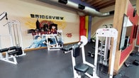Bridgeview Gym is a distinctive fitness facility that blends industrial architectural character with a bold, energetic aesthetic. The space is defined by its soaring exposed wood-beam ceilings supported by heavy timber columns, giving the interior a raw, warehouse-inspired feel. Visible HVAC ductwork runs overhead, reinforcing the industrial character throughout.

The gym floor is covered in durable rubber flooring, providing safe, cushioned surfaces across the entire workout area. The space is divided into functional training zones, each well-equipped for a variety of fitness needs. A comprehensive free weights section offers a full range of dumbbells on tiered racks alongside adjustable benches for pressing and rowing exercises. Multiple strength machines by York USA and Body Solid are stationed throughout, targeting a wide array of muscle groups.

One section of the gym features an eye-catching large-scale graffiti mural branded "Bridgeview," adding a vivid urban art element to the space. Exposed brick walls in another zone contribute texture and warmth, accented by bold yellow and purple painted sections. Large mirrors are mounted along select walls to support training visibility. The open layout allows natural light to filter in from upper-level windows, illuminating the workout areas and enhancing the overall energetic ambiance of the facility.