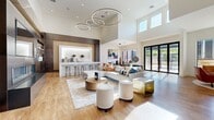 This expansive single-floor amenity facility offers a thoughtfully designed layout across a generous footprint, combining social, wellness, and professional spaces under one roof. The centerpiece is a dramatic open-plan kitchen and living room featuring soaring double-height ceilings, oversized circular pendant lighting, and warm hardwood flooring. A sleek bar-height counter with marble-look stone surfaces, dark cabinetry, and integrated appliances anchors the kitchen area, while the living space accommodates multiple seating arrangements bathed in natural light from large glass doors that open to an outdoor terrace. Adjacent to the main gathering area, a spacious lounge features leather seating, modern pendant fixtures, and expansive windows overlooking the exterior. The dedicated office provides a productive workspace with built-in computer stations, wood flooring, textured accent walls, and ample natural light. The well-appointed fitness center is comprehensively equipped with strength machines, cardio equipment, free weights, stability balls, and ceiling fans, all set on durable carpet tile flooring. Wide, light-filled hallways finished in large-format porcelain tile connect all areas seamlessly, accented by contemporary artwork and clean white walls, creating a polished and welcoming circulation experience throughout.