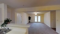 This single-floor space presents an open and functional layout centered around a spacious living area. The room features wall-to-wall carpeting in a neutral tone, contributing to a warm and comfortable atmosphere. A ceiling fan with a light fixture is centrally mounted, providing both air circulation and ambient lighting throughout the space.

Natural light fills the room through a set of double glass doors positioned along the far wall, offering a view of an outdoor area, such as a balcony or patio, and creating a bright, airy feel. The doors serve as a focal point of the living space while enhancing the connection between the interior and exterior.

The walls are painted in soft, neutral tones, complementing the light-colored trim and panel-style interior door visible near the entryway. A textured ceiling adds subtle visual depth to the space. A partial white countertop or bar surface is visible in the foreground, suggesting an adjacent kitchen or transitional area nearby.

Overall, the space is clean, open, and move-in ready, with a straightforward layout that accommodates a variety of furniture arrangements and uses.