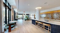 Located at 600 St Albans Dr in Raleigh, NC, this thoughtfully designed amenity floor offers a generous, single-level layout encompassing a variety of distinct functional zones. The entrance greets visitors with large-format terrazzo tile flooring and dramatic floor-to-ceiling black-framed glass doors, setting an immediately refined tone.

The main lounge area features wide-plank herringbone hardwood flooring, coffered ceilings with recessed lighting, and expansive floor-to-ceiling windows that flood the space with natural light and frame sweeping views of the surrounding woodland. Comfortable seating arrangements and curated furnishings create an inviting, residential atmosphere.

A dedicated co-working and library zone offers built-in light wood shelving stocked with books, a backlit counter-height work bar with seating, and intimate arched private booths upholstered in deep navy — ideal for focused work or quiet retreat. An entertainment area features a built-in media wall with flanking shelving and display niches.

The wet bar area is anchored by a striking curved fluted island, warm amber-tiled backsplash, and ample counter space, functioning as a central social hub. Throughout the space, exposed concrete columns, layered area rugs, teal accent walls, and carefully selected artwork contribute to a cohesive, upscale modern aesthetic.