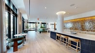 Located at 600 St Albans Dr in Raleigh, NC, this thoughtfully designed amenity floor offers a generous, single-level layout encompassing a variety of distinct functional zones. The entrance greets visitors with large-format terrazzo tile flooring and dramatic floor-to-ceiling black-framed glass doors, setting an immediately refined tone.

The main lounge area features wide-plank herringbone hardwood flooring, coffered ceilings with recessed lighting, and expansive floor-to-ceiling windows that flood the space with natural light and frame sweeping views of the surrounding woodland. Comfortable seating arrangements and curated furnishings create an inviting, residential atmosphere.

A dedicated co-working and library zone offers built-in light wood shelving stocked with books, a backlit counter-height work bar with seating, and intimate arched private booths upholstered in deep navy — ideal for focused work or quiet retreat. An entertainment area features a built-in media wall with flanking shelving and display niches.

The wet bar area is anchored by a striking curved fluted island, warm amber-tiled backsplash, and ample counter space, functioning as a central social hub. Throughout the space, exposed concrete columns, layered area rugs, teal accent walls, and carefully selected artwork contribute to a cohesive, upscale modern aesthetic.