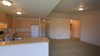 This single-floor unit features an open-concept layout that seamlessly connects the kitchen and living areas, creating a functional and cohesive living space. The kitchen is equipped with light wood cabinetry, a white refrigerator, and a sink, with ample counter space along the perimeter. A peninsula with a light-toned countertop extends outward, providing additional workspace and serving as a natural divider between the kitchen and the adjoining living area.

The living space is finished with wall-to-wall neutral-toned carpeting, lending a comfortable and understated aesthetic throughout. Warm ceiling-mounted light fixtures illuminate the area, complementing the beige and off-white wall tones. Architectural detailing includes subtle ceiling transitions that add dimension to the interior without overwhelming the overall design.

Toward the rear of the space, multiple doorways are visible, suggesting access to additional rooms or storage, with a hallway leading further into the unit. The layout is practical and straightforward, making efficient use of the available footprint. Overall, the space presents a clean, move-in-ready environment with a simple, functional design.