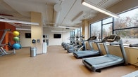 This fitness center is designed to accommodate a comprehensive range of exercise needs within a spacious, open-plan layout. The facility is divided into two primary zones: a cardio area and a strength training area, each thoughtfully outfitted with professional-grade equipment.

The cardio zone features a row of treadmills and elliptical machines positioned along a wall of large, floor-to-ceiling windows. These expansive black-framed windows flood the space with natural light and offer views of surrounding trees and outdoor scenery, creating an energizing atmosphere during workouts. The flooring in this zone is a durable, cushioned material well-suited for high-traffic use, and overhead fluorescent strip lighting supplements the natural light.

The strength training area transitions to a darker rubber floor, providing a sturdy surface for free weights and resistance machines. This zone is equipped with a full dumbbell rack, adjustable weight benches, a power rack, cable machines, and a variety of resistance equipment. A bold orange accent wall adds visual energy to this section, and a wall-mounted flat-screen television is positioned for easy viewing.

Throughout the facility, exposed ductwork runs along the ceiling, contributing to an industrial aesthetic. Additional amenities include a water station, sanitation supplies, and conveniently placed waste receptacles. The overall design balances functionality with an open, airy ambiance suited to a wide range of fitness activities.