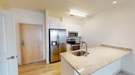 This single-story unit offers a clean, modern aesthetic throughout, highlighted by light wood-style flooring that flows seamlessly across all living areas. The open-concept kitchen serves as a central feature, equipped with stainless steel appliances including a side-by-side refrigerator with ice and water dispenser, an electric range, and a built-in microwave. White cabinetry extends to the ceiling for ample storage, while a peninsula with granite countertops provides additional workspace and a stainless steel undermount sink. The kitchen opens directly into a generously proportioned living area, which benefits from natural light through windows fitted with horizontal blinds and a ceiling fan overhead.

The layout includes a dedicated hallway leading to the private areas of the unit. The bedroom features a ceiling fan with light, a window allowing natural light, and light-toned flooring consistent with the rest of the space. The bathroom includes a full tub-shower combination with brushed nickel fixtures. A notable practical feature is the in-unit laundry area with a stacked washer and dryer. Closet space with wire shelving provides organized storage. The overall ambiance is bright, clean, and move-in ready.
