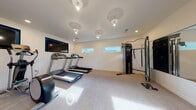 Located at 1821 Brookwood Dr in Vacaville, California, this single-story fitness center is thoughtfully designed to support a comprehensive workout experience. The space is organized across two interconnected areas, each serving distinct exercise functions while maintaining a cohesive, modern aesthetic throughout.

The primary cardio room features neutral-toned carpet flooring and bright white walls, creating a clean and energizing environment. Multiple treadmills are positioned for easy use, accompanied by a wall-mounted flat-screen television for entertainment during workouts. A cable strength training machine and additional resistance equipment occupy one side of the room, while horizontal clerestory windows allow natural light to filter in without compromising wall space for equipment placement. Recessed and pendant lighting fixtures illuminate the room evenly overhead.

The adjoining functional training area introduces a striking geometric-patterned textured accent wall that serves as a bold visual focal point. This room accommodates a stationary bike, elliptical trainer, adjustable weight bench, and a well-organized storage rack holding medicine balls, resistance bands, exercise mats, and stability balls. A wall-mounted water station with a built-in bottle filler adds convenience. A glass door opens directly to an outdoor area, bringing in additional natural light and providing easy access to the surrounding grounds.