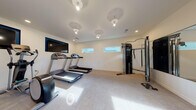 Located at 1821 Brookwood Dr in Vacaville, California, this single-story fitness center is thoughtfully designed to support a comprehensive workout experience. The space is organized across two interconnected areas, each serving distinct exercise functions while maintaining a cohesive, modern aesthetic throughout.

The primary cardio room features neutral-toned carpet flooring and bright white walls, creating a clean and energizing environment. Multiple treadmills are positioned for easy use, accompanied by a wall-mounted flat-screen television for entertainment during workouts. A cable strength training machine and additional resistance equipment occupy one side of the room, while horizontal clerestory windows allow natural light to filter in without compromising wall space for equipment placement. Recessed and pendant lighting fixtures illuminate the room evenly overhead.

The adjoining functional training area introduces a striking geometric-patterned textured accent wall that serves as a bold visual focal point. This room accommodates a stationary bike, elliptical trainer, adjustable weight bench, and a well-organized storage rack holding medicine balls, resistance bands, exercise mats, and stability balls. A wall-mounted water station with a built-in bottle filler adds convenience. A glass door opens directly to an outdoor area, bringing in additional natural light and providing easy access to the surrounding grounds.