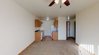 This single-floor unit offers a functional and comfortable layout designed for everyday living. The open-concept living area is finished with plush carpet and features a ceiling fan with light fixture, creating a welcoming atmosphere. Large sliding glass doors dressed with vertical blinds allow generous natural light to fill the space and provide outdoor views. The living area flows seamlessly into the kitchen, which is equipped with warm oak cabinetry, dark granite-style countertops, and a full suite of white appliances including a refrigerator, electric range, dishwasher, and over-the-range microwave. Light wood-style flooring in the kitchen adds a clean, practical contrast to the carpeted living spaces. Two well-sized bedrooms each feature soft carpet, vertical blind-covered windows that invite natural light, and wall-mounted HVAC units for individual climate control. The full bathroom is finished with light vinyl flooring, a single-sink oak vanity with mirrored medicine cabinet, a soaking tub with shower surround, and a chrome towel bar. A hallway connects all areas of the unit, with convenient entry access near the kitchen. The overall aesthetic is clean, neutral, and move-in ready.