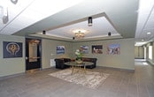 This single-floor commercial space presents a refined and professional environment, thoughtfully designed to combine functionality with a sophisticated aesthetic. The lobby area features large-format light gray tile flooring that extends seamlessly into a wide, well-lit corridor, creating a sense of openness and continuity throughout. A coffered ceiling detail in the lobby adds architectural depth, complemented by a mix of recessed lighting and a decorative chandelier that together create a warm, layered ambiance. The space is adorned with curated artwork, including a prominently displayed framed American flag that serves as a striking focal point, alongside a series of colorful landscape photographs that add character and visual interest to the walls. A classic tufted leather sofa and a round accent table with an area rug define a comfortable seating nook in the lobby, offering a welcoming respite for visitors. A rustic wooden console beneath the flag artwork adds a warm, textural contrast to the otherwise contemporary surroundings. The corridor stretches toward a window-lit end, drawing in natural light and enhancing the airy, open feel of the space. Overall, the design strikes a balance between professional formality and approachable warmth.