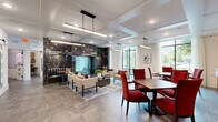 This elegantly appointed amenity lounge spans a single floor and is thoughtfully designed to accommodate a variety of social and recreational needs. The space is anchored by a striking floor-to-ceiling dark marble fireplace surround, which serves as a dramatic architectural focal point and naturally divides the lounge into distinct functional zones.

The main gathering area features a comfortable seating arrangement with neutral-toned sofas and accent chairs set atop a layered area rug, ideal for relaxed conversation. Adjacent to this, a dining area outfitted with rich wood tables and bold red upholstered chairs provides a more structured setting, complemented by a large-screen display and built-in shelving along one wall.

A separate, more intimate nook offers a round wood dining table beside floor-to-ceiling windows, framed by a textured grasscloth accent wall and curated artwork, with direct views of a landscaped outdoor terrace. The entry lounge area features swivel chairs, a tufted velvet bench, and gold-accented side tables arranged around the fireplace, alongside white sliding barn doors that lead to additional private rooms.

Throughout, large-format stone tile flooring, coffered ceilings with linear pendant lighting, and expansive windows flood the space with natural light, creating a polished yet welcoming atmosphere.
