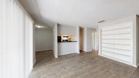 Located at 7600 Blanco Rd in San Antonio, Texas, this single-story unit offers a practical and functional layout designed for comfortable everyday living. The open living area is finished with durable wood-look flooring that extends throughout the space, complemented by neutral gray walls and a textured ceiling. A large sliding glass door floods the living room with natural light and provides views of the surrounding greenery, with vertical blinds offering added privacy.

The kitchen is positioned behind a half-wall pass-through, creating a semi-open connection to the living area. It is outfitted with white cabinetry, dark countertops, and black appliances including an electric range and refrigerator. A window above the kitchen passthrough brings in additional natural light.

A built-in shelving unit near the entryway adds convenient display and storage space. The bedroom is a well-proportioned room featuring wood-look flooring, a ceiling fan with light fixture, and a window with blinds that allow natural light to filter in.

The bathroom includes a white vanity with dark countertops, a framed mirror, and wood-look flooring that maintains consistency with the rest of the unit. A separate laundry area with stacked appliances is accessible from the bathroom corridor, adding to the overall practicality of the space.