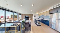 Located in Seattle's South Lake Union neighborhood, this contemporary clubroom offers a well-appointed, open-plan layout that seamlessly integrates a kitchen and living area into a single cohesive space. The room is finished with large-format porcelain floor tiles and recessed ceiling lighting throughout, creating a clean, modern aesthetic.

The kitchen is a functional centerpiece, featuring bold navy-blue lower cabinetry paired with light wood upper cabinets and a soft blue subway tile backsplash. A stainless steel island with bar seating provides an ideal gathering spot, complemented by a full-size stainless steel refrigerator and a matte black sink fixture with white countertops.

The living area is anchored by a striking vertical wood slat accent wall, against which a large flat-screen television is mounted above a geometric-patterned media console. Plush sectional seating, layered area rugs, and curated accent furnishings contribute to a comfortable yet stylish atmosphere.

A defining feature of this space is the floor-to-ceiling sliding glass doors that open directly onto an outdoor terrace, flooding the interior with natural light and offering panoramic views of the Seattle skyline. The terrace features raised planters with greenery, extending the room's functionality outdoors and enhancing the overall sense of openness.