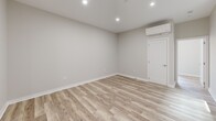 This thoughtfully renovated one-bedroom, one-bathroom unit offers a clean, modern aesthetic across a single floor. Light-toned hardwood flooring flows throughout the main living areas, complementing the neutral gray-beige walls and recessed lighting that create a bright, airy atmosphere. The living room features a wall-mounted mini-split HVAC unit and large black-framed windows that invite natural light and offer views of the surrounding urban streetscape. The well-appointed kitchen is a standout, combining white upper shaker cabinets with gray lower cabinets, crisp white quartz countertops, a large center island, and stainless steel appliances including a gas range, microwave, and French door refrigerator. An adjacent utility kitchen area showcases an exposed white-painted brick wall, gray tile flooring, a stainless steel sink, and additional cabinetry, adding both character and practicality. The spacious primary bedroom includes a double-door closet, wall-mounted HVAC, and recessed lighting. The bathroom is finished with marble-style wall tile, gray floor tile, a gray vanity with a white countertop, and a backlit LED mirror. A utility room rounds out the functional layout of this move-in-ready space.