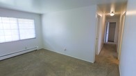 This single-floor residential space offers a clean, functional layout with a simple and practical design throughout. The interior is characterized by bright white walls that create a light, open atmosphere, complemented by wall-to-wall carpeting that adds warmth and comfort underfoot. A window allows natural light to filter into the main living area, contributing to the airy feel of the space. A baseboard heating unit is present along the wall, providing an efficient source of warmth. The room opens through a doorway that leads further into the rest of the space, suggesting a connected and accessible floor plan. The neutral color palette and understated finishes lend the interior a versatile quality, making it easy to personalize. Overall, the space presents a straightforward, move-in ready environment suited for everyday residential living.