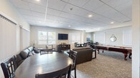 Located at 550 Park Ave W in Chatham-Kent, Ontario, this single-floor communal space offers a thoughtfully designed layout that combines a living room, dining area, and kitchen into one cohesive and functional environment. The living room is comfortably furnished with leather sofas, an accent chair, and a central coffee table, all arranged around a wall-mounted flat-screen television. Large windows fitted with vertical blinds allow natural light to fill the space while providing outdoor views of the surrounding area. A drop ceiling with recessed lighting contributes to the room's clean, contemporary aesthetic. The dining area accommodates multiple round tables with dark wood chairs, making it well-suited for group gatherings. Natural light streams through additional windows, brightening the patterned carpet flooring. A chalkboard and indoor greenery add character to the dining nook. A pool table is also present, providing an additional recreational feature. The kitchen is efficiently designed with rich dark wood cabinetry, light granite-style countertops, and a classic subway tile backsplash. It is equipped with a full-size stainless steel refrigerator, a microwave, and a double sink, all set atop light-toned tile flooring. Together, these areas form a versatile and welcoming space ideal for community use.