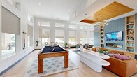 This expansive single-floor amenity space offers a generous, open-concept layout designed for both socializing and relaxation. Soaring double-height ceilings flood the interior with natural light through rows of large clerestory windows, creating an airy, inviting atmosphere throughout.

The main living area serves as the focal point, anchored by a striking feature wall clad in blue-grey tile, built-in shelving, and a sleek fireplace topped by a wall-mounted television. A warm wood-paneled tray ceiling with suspended circular gold pendant lights adds elegance and visual contrast overhead. Comfortable, eclectic seating arrangements — including teal armchairs, leather sofas, and ottomans — invite gathering and conversation.

Adjacent to the living area, a dedicated games zone houses a full-size billiards table set atop a geometric area rug, with cue racks mounted nearby for easy access. A cozy dining nook features built-in wood-paneled banquette seating with colorful cushions, paired with a round table and modern chairs.

The kitchen area is outfitted with an expansive quartz island, navy cabinetry with brass hardware, beverage refrigerators, and a wet bar with matte black fixtures — all beneath dramatic woven pendant lights. Light wood herringbone-pattern flooring flows seamlessly throughout, tying the entire space together with a contemporary yet warm aesthetic.
