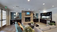 This single-floor communal lounge space is thoughtfully designed to provide a warm and inviting atmosphere for residents and guests alike. The room's centerpiece is a striking floor-to-ceiling stacked stone fireplace, topped with a classic mantel and a wall-mounted flat-screen TV, creating an immediate focal point that anchors the entire space. Recessed lighting throughout the ceiling provides consistent, ambient illumination, complementing the light wood-toned flooring that runs across the room. The walls are finished in a rich teal accent color, adding depth and a modern touch to the overall aesthetic. Built-in dark wood cabinetry lines both sides of the fireplace wall, offering display shelving and storage, while decorative framed mirrors add visual interest and a sense of openness. A well-appointed seating arrangement features plush upholstered sofas and teal accent chairs with nailhead trim, arranged around a central coffee table atop a patterned area rug. Lush greenery and decorative accents complete the décor. To one side, a small refreshment station with countertop appliances is conveniently integrated into the cabinetry, making this space ideal for casual socializing and relaxation.