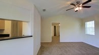 This single-floor home offers a clean, functional layout with several notable architectural details. The living area is highlighted by vaulted ceilings that lend a sense of openness and height, complemented by a ceiling fan that enhances both comfort and airflow. Soft, wall-to-wall carpeting runs throughout the living space, creating a warm and inviting atmosphere underfoot.

A built-in pass-through counter connects the living area to the kitchen, providing a practical and convenient interface between the two spaces. The kitchen counter features a dark countertop, offering a subtle contrast to the light-toned walls that are consistent throughout the home.

The neutral white walls and textured ceiling contribute to a bright and airy feel, while a centrally positioned HVAC vent ensures consistent climate control. A doorway from the living area leads into a hallway that connects to additional rooms, suggesting a practical and well-organized floor plan.

Overall, the space presents a straightforward and comfortable interior, characterized by its vaulted ceilings, open sightlines, and cohesive neutral color palette — making it an adaptable canvas for a variety of furnishing and décor styles.