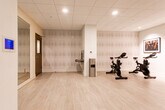 Located at 1 Woodbridge Center Drive in Woodbridge, NJ, this modern fitness studio offers a thoughtfully designed, single-floor layout ideal for a variety of workout disciplines. The space features light hardwood-style flooring throughout, a drop tile ceiling with recessed lighting, and a clean, neutral color palette of whites and soft grays that creates a bright and energizing atmosphere.

The main studio area is equipped with multiple exercise mats and is lined with large floor-to-ceiling mirrors that visually expand the space and allow for proper form monitoring during workouts. A wall-mounted flat-screen television serves as a focal point, making the room well-suited for guided fitness classes or virtual instruction. Camera tripods are also present, indicating the space supports recorded or live-streamed sessions.

A dedicated cycling area houses high-quality stationary spin bikes positioned in front of a textured, vertically striped accent wall, adding depth and visual interest to the room. Framed artwork depicting energetic, music-inspired photography adorns the walls throughout, lending the studio a polished, gallery-like feel.

A glass partition wall with a dark-framed door separates areas within the facility, providing both visibility and privacy as needed. A water station and wall-mounted thermostat ensure occupant comfort. The overall design balances functionality with a refined aesthetic, making this a professional and welcoming environment for fitness and wellness activities.