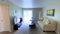 This single-floor space offers a comfortable and well-lit living environment designed for everyday functionality. The living room serves as the centerpiece of the layout, featuring wall-to-wall carpeting that adds warmth and softness underfoot. A large sliding glass door dressed with vertical blinds dominates one wall, allowing generous amounts of natural light to pour in and providing a view to the outdoors. The room is finished with a textured ceiling, adding a subtle architectural detail overhead. Neutral-toned walls create a clean, versatile backdrop that complements a variety of furnishing styles. The space comfortably accommodates a full seating arrangement, with room for a sofa, accent chair, coffee table, and an entertainment center. A tall floor lamp in the corner adds ambient lighting options. The overall ambiance is calm and inviting, with the natural light from the sliding door being a standout feature that enhances the sense of openness throughout the living area. The simple, understated design makes this space highly adaptable to personalization while maintaining a functional and welcoming atmosphere.
