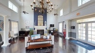This single-story clubhouse makes a striking first impression with its soaring double-height ceilings and dramatic grand chandelier, which anchors the main gathering area with timeless elegance. Ceiling fans complement the open layout, ensuring comfort throughout the space. The focal point of the main lounge is a classic white-mantled fireplace set against a bold navy blue accent wall, topped with a large arched mirror that amplifies the sense of height and light. Plush white sofas and a wood coffee table create an inviting seating arrangement, ideal for casual socializing or community gatherings. Dark polished tile floors run throughout, lending a sophisticated, cohesive look to the interior. A dining area with wooden chairs is positioned near windows that allow generous natural light to fill the room. A dedicated "Success Station" with desk space provides a functional workspace for residents. Additional amenities include a classic red popcorn machine, adding a fun and welcoming touch. Decorative columns and neutral wall tones balance the bold design elements, resulting in an atmosphere that is both refined and approachable — a true community hub designed for everyday enjoyment.