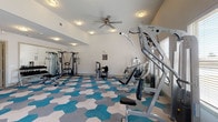 This fitness center is designed to support a comprehensive workout experience within a single, open-plan space. The room is generously proportioned, offering ample floor space to accommodate a diverse array of exercise equipment without feeling crowded. A striking hexagonal tile pattern in shades of teal, gray, and cream covers the entire floor, giving the space a modern, energetic aesthetic that sets it apart from a standard gym environment.

The cardio section lines one wall, featuring multiple treadmills, elliptical trainers, and stationary bikes positioned in front of large windows fitted with blinds. These windows allow substantial natural light to fill the room while providing views of the surrounding exterior. The strength training area occupies the remaining floor space, equipped with a variety of cable machines, weight stations, and functional training equipment to target multiple muscle groups.

Overhead, flush-mounted ceiling lights and a ceiling fan ensure the space remains well-lit and well-ventilated. A built-in counter with shelving and a bold orange accent wall in an adjoining entryway area provides a functional transition zone into the fitness floor. The clean white walls and neutral tones throughout contribute to a bright, focused atmosphere conducive to exercise.