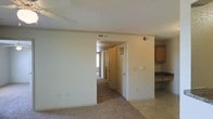 This single-floor residential space offers a straightforward and functional layout designed for comfortable everyday living. The main living area features wall-to-wall carpeting in a neutral tone, creating a warm and cohesive feel throughout. A textured ceiling adds a classic residential character to the space, while multiple HVAC vents ensure consistent climate control throughout the unit.

An open archway connects the living area to a hallway that leads to additional rooms, providing a natural flow between spaces. The hallway is well-lit by a window at its far end, allowing natural light to filter into the interior. Doors along the hallway suggest access to private rooms or storage areas.

To one side, a kitchen area is partially visible, featuring dark wood cabinetry and a countertop, offering practical storage and workspace. The kitchen's placement adjacent to the main living area supports an efficient and connected floor plan.

The overall ambiance of the space is clean, neutral, and move-in ready, with light-toned walls throughout that lend an open and airy feel. The consistent use of carpet, neutral paint, and simple finishes gives the space a cohesive and understated aesthetic suitable for a variety of furnishing styles.