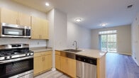 This well-appointed single-floor residence offers a clean, modern aesthetic throughout its thoughtfully designed layout. The open-concept kitchen serves as a central hub, featuring light maple cabinetry, granite-style countertops, and a full suite of stainless steel appliances including a gas range, built-in microwave, refrigerator, and dishwasher. A peninsula island with an undermount double sink adds both functionality and a natural division between the kitchen and the open living area. Recessed lighting illuminates the kitchen, complementing the light wood-style flooring that flows seamlessly through the main living spaces. The living area is bright and open, benefiting from a sliding glass door that leads to a private balcony with views of surrounding greenery, allowing natural light to fill the space. The bedrooms offer plush carpeted flooring and window access to natural light, providing a comfortable and quiet retreat. The bathroom is finished with a wood-toned vanity, granite-style countertop, an undermount sink, and a large mirror, all set against neutral-toned walls and matching wood-style flooring. Clean white walls and neutral tones throughout lend the space a bright, move-in-ready appeal.