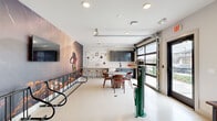 Located at 5000 Collina Ter in Clermont, FL, this single-floor community amenity space is thoughtfully designed around an active, outdoor lifestyle. The open-plan layout is anchored by a dramatic floor-to-ceiling mountain biking mural that commands immediate attention, setting an energetic and adventurous tone throughout the room. Polished concrete-style flooring flows seamlessly across the entire space, lending a clean, modern aesthetic that is both durable and visually cohesive.

At the heart of the room sits a round dining table surrounded by warm wood-toned chairs, perfect for casual gatherings. Adjacent to it, a butcher block-topped counter with bar seating extends along the wall, complemented by sleek navy cabinetry and a pegboard organizer — ideal for communal use. A dedicated bike repair station with a professional-grade floor pump stands ready near the glass roll-up garage door, which floods the space with natural light and provides easy outdoor access.

A quieter lounge nook at one end offers a plush green sofa, a framed cycling-themed art print, and a wall-mounted mini-split for climate control, creating a relaxed retreat within the larger space. A wall-mounted television near the mural adds an entertainment element. This space is a well-equipped hub for cycling enthusiasts and community members alike.