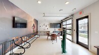 Located at 5000 Collina Ter in Clermont, FL, this single-floor community amenity space is thoughtfully designed around an active, outdoor lifestyle. The open-plan layout is anchored by a dramatic floor-to-ceiling mountain biking mural that commands immediate attention, setting an energetic and adventurous tone throughout the room. Polished concrete-style flooring flows seamlessly across the entire space, lending a clean, modern aesthetic that is both durable and visually cohesive.

At the heart of the room sits a round dining table surrounded by warm wood-toned chairs, perfect for casual gatherings. Adjacent to it, a butcher block-topped counter with bar seating extends along the wall, complemented by sleek navy cabinetry and a pegboard organizer — ideal for communal use. A dedicated bike repair station with a professional-grade floor pump stands ready near the glass roll-up garage door, which floods the space with natural light and provides easy outdoor access.

A quieter lounge nook at one end offers a plush green sofa, a framed cycling-themed art print, and a wall-mounted mini-split for climate control, creating a relaxed retreat within the larger space. A wall-mounted television near the mural adds an entertainment element. This space is a well-equipped hub for cycling enthusiasts and community members alike.