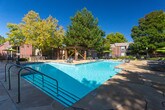 Located at 5801 Eubank Blvd NE in Albuquerque, New Mexico, this single-level outdoor amenity space centers around a generously sized swimming pool with crystal-clear turquoise water. The pool is bordered by a wide concrete deck outfitted with lounge chairs and shaded umbrella seating, providing ample room for relaxation under the abundant New Mexico sun. A prominent wooden pergola structure anchors one end of the pool area, offering a shaded gathering spot with additional seating and a raised deck platform. Adjacent to the pergola, a grill station and landscaped garden beds with ornamental shrubs and gravel accents create a well-appointed outdoor entertaining area. Tall mature trees, including pine and deciduous varieties, frame the perimeter of the space, providing natural shade and privacy. The surrounding buildings feature classic brick and stucco exteriors, with large windows that overlook the pool area. A wrought iron fence encloses the grounds, and manicured green lawn areas are visible beyond the pool deck. The stonework flooring near the entry features flagstone-style paving with a brick accent wall, lending a welcoming, polished finish to the entrance of this outdoor recreational space.