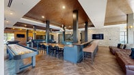 Located at 6408 Providence Farm Lane in Charlotte, NC, this generously sized single-floor community clubhouse offers a dynamic and well-appointed social hub. The open-concept layout flows seamlessly across distinct activity zones, all unified by richly textured herringbone-patterned flooring and dramatic coffered ceilings finished in warm copper and deep wood tones. Bold dark structural columns anchor the space visually while defining each area without sacrificing openness.

A full-length bar with high-backed upholstered seating serves as a central gathering point, complemented by café-style tables and industrial-chic chairs beneath a cluster of globe pendant lights. Exposed brick accent walls lend rustic warmth throughout, contrasting beautifully with the modern finishes. The lounge zone features plush leather sofas and tufted ottomans arranged around a wall-mounted display screen, creating an inviting retreat for relaxation.

The game area is well-equipped with a billiards table, shuffleboard, and additional arcade-style games, all illuminated by dramatic recessed and decorative lighting. Private pod-style enclosures offer quieter spaces for focused work or conversation. Large black-framed windows and expansive sliding glass doors flood the interior with natural light and open directly to a landscaped outdoor patio, extending the social atmosphere seamlessly to the exterior.
