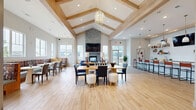 Located at 24 Bay Front Dr in Traverse City, Michigan, this single-floor community clubhouse is thoughtfully designed to serve as a welcoming social hub. The centerpiece of the main gathering area is a dramatic floor-to-ceiling stacked-stone fireplace, topped with a mounted flat-screen TV and flanked by large windows that invite generous natural light. Soaring vaulted ceilings with exposed light wood beam detailing and a striking chandelier lend the space an airy, upscale character. Light hardwood-style flooring flows throughout, unifying the open layout. Multiple seating arrangements accommodate a variety of uses, from casual lounging on plush sofas to more structured dining at large communal tables with mixed seating. A built-in banquette dining nook along a window wall offers an intimate setting for smaller gatherings, bathed in natural light with views of the surrounding greenery. The kitchenette bar area features a clean white shiplap backsplash, open floating shelves with decorative accents, pendant lighting, and counter seating with leather barstools. A dedicated reception or leasing office area includes a sleek white desk, shiplap wall detail, and a textured accent wall. Direct access to an outdoor patio area is available through glass doors adjacent to the fireplace lounge. The overall ambiance is modern, bright, and inviting.
