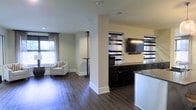 This single-floor space is designed with an open-concept layout that promotes a sense of flow and connectivity between functional areas. Dark hardwood flooring runs throughout, providing a warm, grounding contrast to the light-toned walls. A structural column subtly divides the space without fully closing it off, maintaining visual openness while defining distinct zones.

The lounge area features a cushioned armchair with nailhead trim, positioned near a large window dressed with sheer curtains that allow generous natural light to filter in. A tall accent table accompanies the seating, making the area suitable for casual use or quiet relaxation.

On the opposite side, a built-in cabinetry unit with dark espresso-toned drawers and cabinets provides substantial storage. A wall-mounted flat-screen television is centrally positioned above, flanked by striped horizontal window treatments that add a layered, decorative element. The countertop surface, topped with a stone-look finish, extends along the wall, offering functional workspace. White cabinetry to the right adds contrast and additional storage capacity. The overall ambiance is clean, functional, and contemporary.