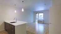 This well-designed single-floor space offers a seamless open-concept layout that effortlessly connects the kitchen and living areas. The kitchen features a clean white countertop island with an undermount sink and contemporary pendant lighting, providing both functional workspace and a stylish focal point. Dark cabinetry contrasts elegantly with the bright white surfaces, lending a modern aesthetic to the space. The living area is bathed in natural light streaming through a large floor-to-ceiling window, which frames a lush outdoor view and creates an inviting, airy atmosphere. Light-toned hardwood floors extend throughout, enhancing the sense of openness and warmth. Crisp white walls and a smooth white ceiling contribute to the clean, minimalist character of the space. A wall-mounted heating unit beneath the window ensures year-round comfort. Recessed lighting complements the pendant fixtures, providing versatile illumination options. The overall ambiance is bright, modern, and welcoming, making it a highly functional and visually cohesive living environment.