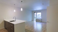 This well-designed single-floor space offers a seamless open-concept layout that effortlessly connects the kitchen and living areas. The kitchen features a clean white countertop island with an undermount sink and contemporary pendant lighting, providing both functional workspace and a stylish focal point. Dark cabinetry contrasts elegantly with the bright white surfaces, lending a modern aesthetic to the space. The living area is bathed in natural light streaming through a large floor-to-ceiling window, which frames a lush outdoor view and creates an inviting, airy atmosphere. Light-toned hardwood floors extend throughout, enhancing the sense of openness and warmth. Crisp white walls and a smooth white ceiling contribute to the clean, minimalist character of the space. A wall-mounted heating unit beneath the window ensures year-round comfort. Recessed lighting complements the pendant fixtures, providing versatile illumination options. The overall ambiance is bright, modern, and welcoming, making it a highly functional and visually cohesive living environment.