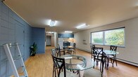 This single-floor communal space is designed to serve as a shared gathering and dining area, combining an open common room with a functional kitchen. The layout is generous and open, featuring warm wood-look flooring throughout that lends a clean, welcoming feel to the interior.

The common room area is furnished with multiple round glass-top dining tables and metal chairs, providing ample seating for group use. Large windows along the exterior walls flood the space with natural light and offer pleasant views of the surrounding greenery. A wall-mounted flat-screen television is positioned for easy viewing across the room. A glass door provides direct access to an outdoor area, where outdoor seating can be glimpsed beyond.

The kitchen is located at one end of the space and is equipped with blue cabinetry — both upper and lower — paired with dark countertops. Appliances include a full-size white refrigerator, a gas range with overhead ventilation, and a sink. Framed artwork decorates the adjacent cinder block wall, adding a touch of warmth to the utilitarian setting.

The walls throughout are painted cinder block, contributing to the space's durable, institutional character. Fluorescent lighting fixtures illuminate the interior evenly. A blue-painted block wall near the entrance features two doors, likely leading to storage or utility areas. Baseboard heating runs along the perimeter, ensuring comfort year-round.