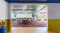Located at 2000 Strand Way in Lancaster, PA, this single-floor amenity space spans an expansive footprint across six distinct areas, offering a comprehensive suite of community features. The interior opens into a sophisticated lounge anchored by a striking green-painted brick fireplace wall with a linear gas fireplace, tufted leather seating, and a coffered ceiling with tray detailing — all contributing to a warm yet modern ambiance. Sliding barn doors add a charming transitional touch between spaces. The adjacent dining and gathering area continues the coffered ceiling motif, now finished in teal, and is filled with natural light from large windows overlooking the outdoors. A built-in refreshment bar and bold, patterned carpeting give this room a vibrant, welcoming character. A dedicated conference or meeting room features deep navy shiplap walls, large picture windows framing lush greenery, and patterned tile flooring for a refined, focused atmosphere. The fitness center is well-equipped with cardio machines, free weights, stability equipment, floor-to-ceiling mirrors, and a bold hexagonal tile floor. Outdoors, a generous resort-style pool is surrounded by a herringbone brick deck lined with white lounge chairs, string lights overhead, and a manicured lawn with shaded seating — perfect for relaxation and socializing.