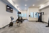 Located at 9815 Almeda Genoa Rd in Houston, Texas, this fitness center offers a well-equipped, single-floor workout space designed to support a comprehensive exercise routine. The room features neutral white and light gray walls accented by a subtle blue band near the ceiling, creating a clean and modern aesthetic throughout. Underfoot, durable patterned carpet tile covers the entire floor, providing a practical and comfortable surface for a variety of workouts. Recessed lighting illuminates the space evenly, while multiple windows fitted with horizontal blinds allow natural light to filter in and offer views of the surrounding exterior. The cardio zone lines one side of the room with a row of treadmills, ellipticals, and a stationary bike, each positioned in front of wall-mounted flat-screen TVs for an engaging workout experience. On the opposite side, a multi-function cable machine and a power rack with a weight bench cater to strength and resistance training needs. A dedicated free weights area features a tiered dumbbell rack alongside a weight bench, offering options for targeted muscle training. The overall layout is thoughtfully organized to maximize the available floor area, ensuring clear circulation between equipment zones. A private entry door provides convenient access, completing this functional and well-appointed fitness facility.