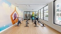This well-appointed fitness studio offers a bright, contemporary space designed to support a comprehensive workout experience. The room is defined by expansive floor-to-ceiling glass windows along its exterior walls, flooding the interior with abundant natural light and providing open views of a resort-style pool area and lush tropical greenery beyond. Light hardwood-style flooring runs throughout, contributing to the clean, energetic atmosphere.

The studio is equipped with a row of Schwinn spin bikes arranged along the windowed wall, ideal for cycling workouts with a view. A rowing machine provides additional cardio options, while a colorful rack of stability balls in varying sizes adds versatility for core and flexibility training. Resistance bands and other functional training accessories are mounted on the wall for easy access.

A glass-panel partition with a large sliding door separates the spin studio from an adjacent fitness area visible through the glass, which houses treadmills and additional strength equipment. A wall-mounted flat-screen television is positioned at the end of the room for entertainment during workouts. One wall features a vibrant motivational mural depicting a yoga pose, adding an artistic and energizing touch to the space. The crisp white ceiling and walls keep the environment feeling open, clean, and modern throughout.