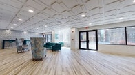 Located in Hartford, CT, this single-floor commercial space offers a thoughtfully designed interior that blends contemporary aesthetics with functional layout. The open floor plan is anchored by light wood-look flooring that runs throughout, creating a clean and cohesive visual foundation. A standout architectural feature is the distinctive geometric coffered ceiling with recessed lighting, adding depth and a refined character to the space.

The main lounge area is furnished with plush teal velvet seating, including a curved sofa and accent chairs, complemented by patterned wingback chairs that introduce a touch of eclectic style. Large street-facing windows flood the interior with natural light and provide clear views of the surrounding Hartford streetscape, reinforcing the space's strong connection to its urban environment. A textured accent wall serves as a striking focal point behind the seating area.

A dedicated bar counter section features a long ledge with bar stools and wall-mounted artwork in teal tones, making it suitable for casual gatherings or light refreshments. The space also includes a reception or service counter area, pointing to its professional, client-facing function. Double glass entry doors at the front provide a welcoming and accessible entry point. Overall, the space presents a polished, modern environment well-suited for professional or community use.