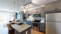 Located at 1104 E Belt Line Rd in Carrollton, TX, this single-story unit offers a thoughtfully designed open-concept layout with modern finishes throughout. The kitchen serves as a central highlight, featuring gray two-tone cabinetry, granite countertops, light blue subway tile backsplash, and a full suite of stainless steel appliances including a refrigerator, range, and microwave. A granite-topped island with pendant lighting above provides additional prep space and casual seating, while track lighting adds functional illumination across the cooking area. Warm wood-look flooring flows throughout the main living spaces, creating a cohesive and inviting atmosphere. The open layout connects the kitchen seamlessly to the living area, which features a teal accent wall and access to adjacent rooms. The bedroom is carpeted and receives natural light through a generously sized window. The bathroom is finished with large-format tile flooring, a granite vanity with dark cabinetry, a wide frameless mirror, and a tub-shower combination. The overall design balances contemporary aesthetics with practical functionality, presenting a clean, modern living environment in a well-located area of Carrollton.
