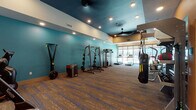 This fitness center is a thoughtfully designed, single-floor facility that offers a comprehensive workout environment for users of all training styles. The space is visually defined by bold teal accent walls paired with warm orange highlights, creating an energetic and motivating atmosphere throughout. A dark, recessed ceiling with recessed lighting and ceiling fans adds depth and contrast to the open layout, while patterned, multi-tone carpet tiles provide a durable and comfortable underfoot surface across the entire floor.

The strength training zone is generously outfitted with a power rack loaded with weight plates, a TRX suspension system, resistance cable machines, a TRX/TRX-style functional trainer, an Everlast heavy punching bag, plyo boxes, and a full rack of dumbbells arranged along a mirrored wall — ideal for free weight and functional training exercises. A stationary bike by Vision Fitness anchors the cardio side, complemented by multiple treadmills and elliptical machines positioned along a wall of expansive, floor-to-ceiling windows. These large windows allow abundant natural light to fill the space and offer open views of the surrounding outdoor courtyard and building exterior. The mirrored wall in the free weights area further enhances the sense of spaciousness, making this fitness center a fully functional and visually inviting training destination.