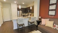 This single-story space showcases an open-concept layout that connects the kitchen and living area, creating a cohesive and functional environment. The kitchen is outfitted with dark espresso cabinetry that contrasts elegantly against light granite countertops and stainless steel appliances, including a full-size refrigerator and built-in microwave. A peninsula-style breakfast bar extends from the kitchen, providing casual seating for two with stylish nailhead-trim bar stools. Pendant lighting hangs above the bar, adding focused illumination and a decorative touch. Dark hardwood-style flooring runs throughout, grounding the space with a warm, contemporary feel. An accent wall in a rich terracotta hue anchors the living area, adding depth and visual interest alongside a neutral-toned sofa. Recessed lighting in the ceiling complements the natural and artificial light sources throughout. The overall design balances bold color choices with clean, modern finishes, resulting in a space that feels both inviting and well-appointed.