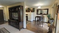 This single-floor space offers an efficient and well-appointed open-concept layout that connects the living, dining, and kitchen areas in a cohesive and functional arrangement. The living area features plush carpeting and is anchored by a dark-stained wooden entertainment center, providing ample storage and a focal display space. Transitioning toward the interior, rich hardwood-style flooring runs through the dining and kitchen zones, adding warmth and visual continuity throughout. The dining nook is furnished with a bistro-style bar table and matching dark wood chairs, complemented by a large framed mirror on the wall that enhances the sense of space and light. The kitchen is equipped with dark cabinetry, a white refrigerator, and stone-look countertops, with a decorative area rug adding a touch of character to the tiled floor. Greenery accents are thoughtfully placed atop the cabinetry, bringing a natural element into the space. Flush-mount ceiling fixtures provide consistent lighting throughout. The cohesive use of dark wood tones across the furniture, cabinetry, and décor creates a unified, warm aesthetic that makes this space feel both stylish and inviting.