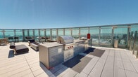 Perched high above Long Beach at 777 E Ocean Blvd, this expansive rooftop amenity deck offers an exceptional open-air experience with sweeping panoramic views of the city skyline, harbor, and surrounding coastline. Enclosed by floor-to-ceiling glass panel railings, the space maximizes visibility while providing a sense of openness under the Southern California sky.

At the heart of the rooftop is a resort-style swimming pool finished with a wood-plank deck surround and lined with rows of cushioned lounge chairs, ideal for sunbathing with unobstructed views of the horizon. Adjacent to the pool, a fully equipped outdoor kitchen serves as a premier entertaining hub, featuring a stainless steel built-in grill, a secondary cooking surface, and tile-clad cabinetry — all designed for functionality and style.

A dedicated lounge area showcases a concrete fire pit table flanked by contemporary wood-framed benches with dark cushioned seating, creating an inviting gathering space. A high-top bistro table with wicker chairs is positioned along the glass perimeter, offering a more intimate vantage point over the cityscape below.

The modern glass curtain wall of the building serves as a striking architectural backdrop throughout the deck, reflecting the sky and reinforcing the elevated, urban character of this rooftop retreat.