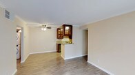 Located at 5427 Lake Murray Boulevard in La Mesa, this single-story unit offers a clean, open layout designed for comfortable everyday living. The main living area is spacious and bright, finished with wood-style laminate flooring that runs seamlessly throughout the space. Crown molding lines the ceiling, adding a refined architectural detail, while a ceiling fan with an integrated light fixture provides both comfort and ambient lighting.

The kitchen is positioned at the rear of the living area and features rich, dark wood cabinetry with glass-front upper cabinets that lend a classic, warm aesthetic. A peninsula with a light-toned granite or stone countertop separates the kitchen from the living space, functioning as a breakfast bar and providing additional counter workspace. A stainless steel appliance is visible beneath the counter, contributing to the kitchen's functional design. The neutral, warm-toned walls throughout complement the wood finishes and create a cohesive, inviting atmosphere. The open flow between the living and kitchen areas makes the space feel connected and well-suited for both daily use and casual entertaining.
