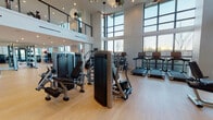 This contemporary fitness center is defined by its dramatic double-height ceilings and expansive floor-to-ceiling windows that flood the space with natural light and frame outdoor views. Light hardwood flooring runs throughout, contributing to a clean, modern aesthetic that pairs seamlessly with white walls and recessed lighting.

The main floor is thoughtfully organized into distinct training zones. A dedicated strength area features a comprehensive selection of resistance machines, free weights, dumbbells, kettlebells, and adjustable benches, with full-length mirrors along the walls to enhance both form and spatial depth. A cardio zone houses treadmills, ellipticals, stationary bikes, and a stair climber, positioned to take advantage of the natural light streaming through the large windows.

A striking steel-and-wood staircase with cable railings connects to the mezzanine level above, which accommodates additional cardio equipment and cycling bikes beneath sculptural pendant lighting. Built-in cubbies beneath the staircase provide organized storage for yoga mats and accessories. The entry is marked by elegant navy-framed glass doors, adding an architectural focal point. Overall, the space delivers a refined, functional, and energizing environment for a comprehensive fitness experience.
